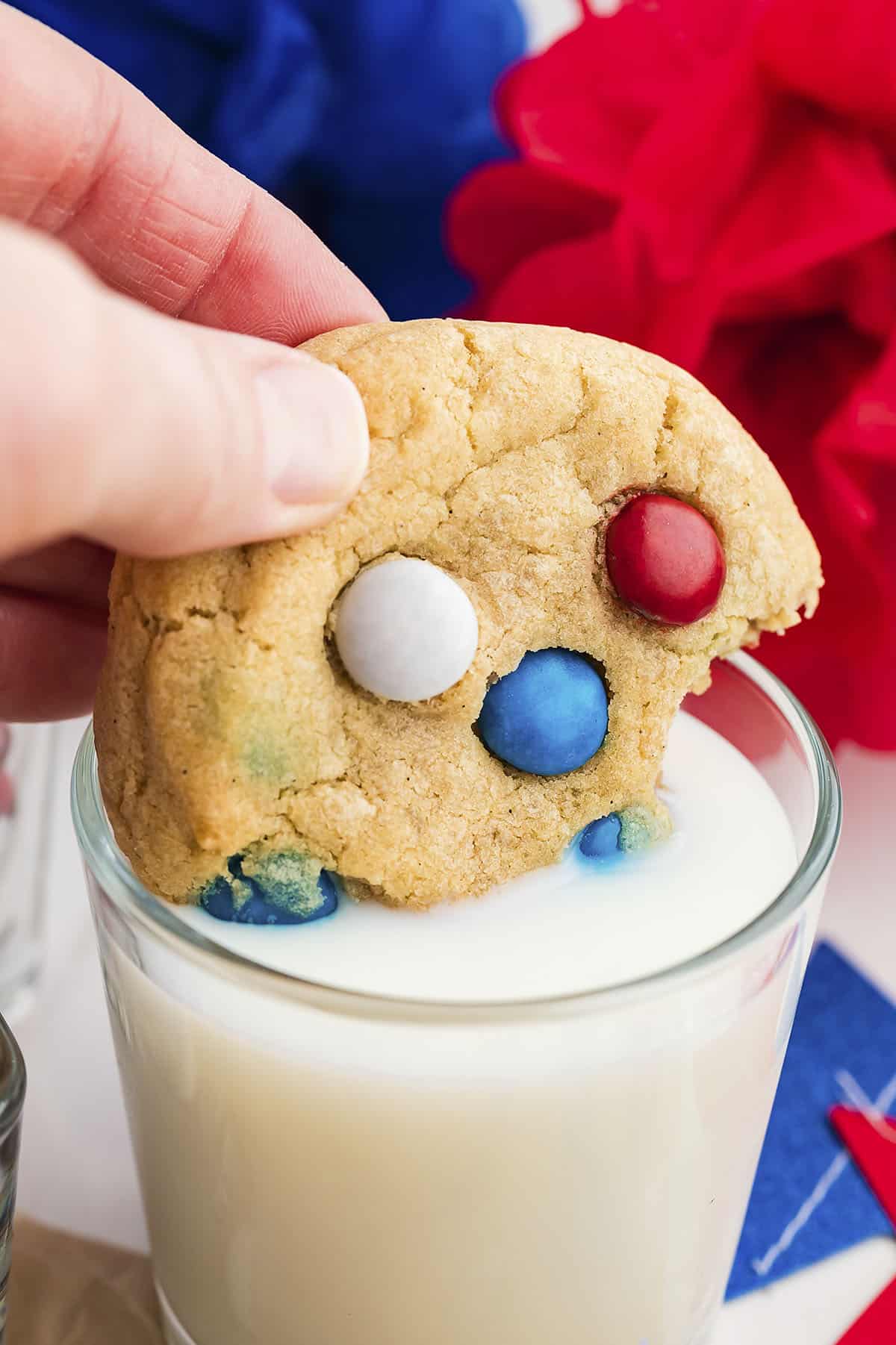 Hand dipping a 4th of July cookie with m&ms into a glass of milk.