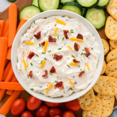 Cheddar bacon ranch dip in bowl on board with crackers and vegetables.